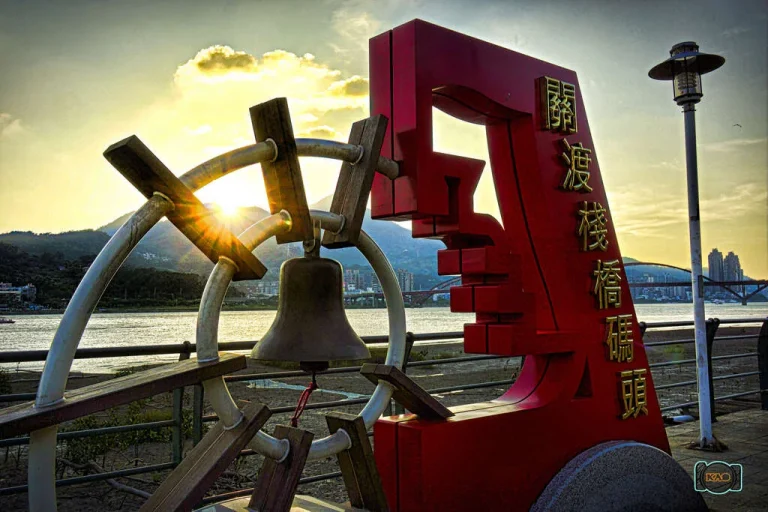 關渡碼頭，夕照下舵輪造型裝置與紅色碼頭地標矗立河岸欄杆旁