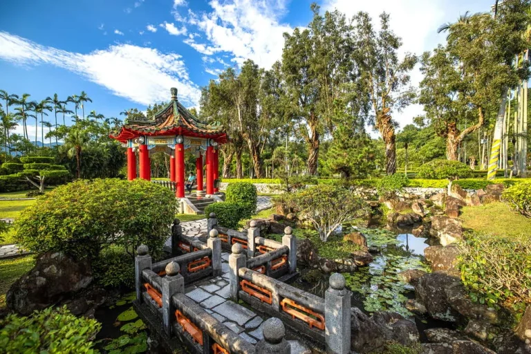 士林官邸公園，紅柱涼亭旁石橋跨越荷花池，藍天下椰影與綠樹相映