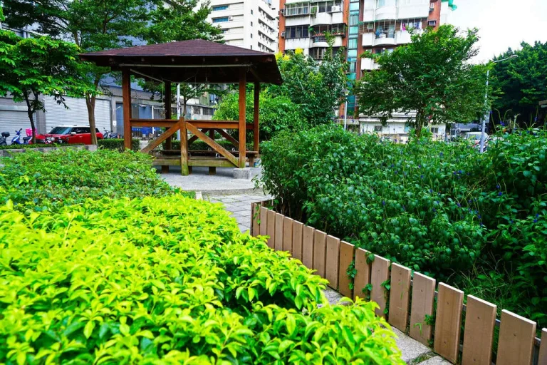 臺北市松錦公園，木造涼亭旁嫩綠灌木叢與木柵欄，後方住宅樓房