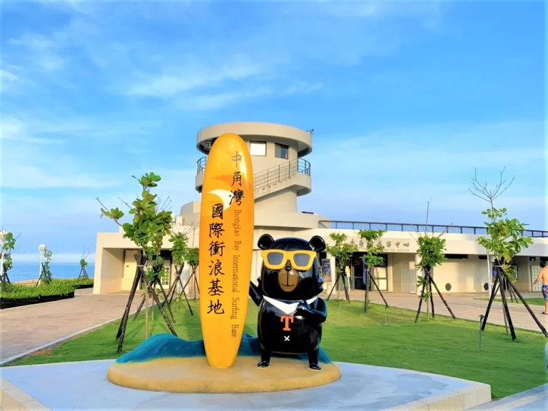 中角灣，黑熊衝浪客公仔與黃色衝浪板地標立於遊客中心前廣場，藍天遼闊
